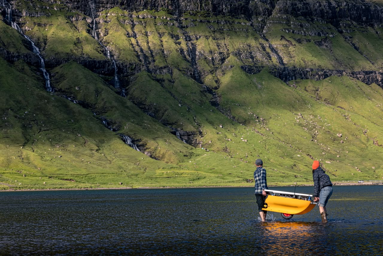 Two BIUTOPIC team members carry HYDRO-01 into a Faroe Islands fjord, waterfall-streaked cliffs behind