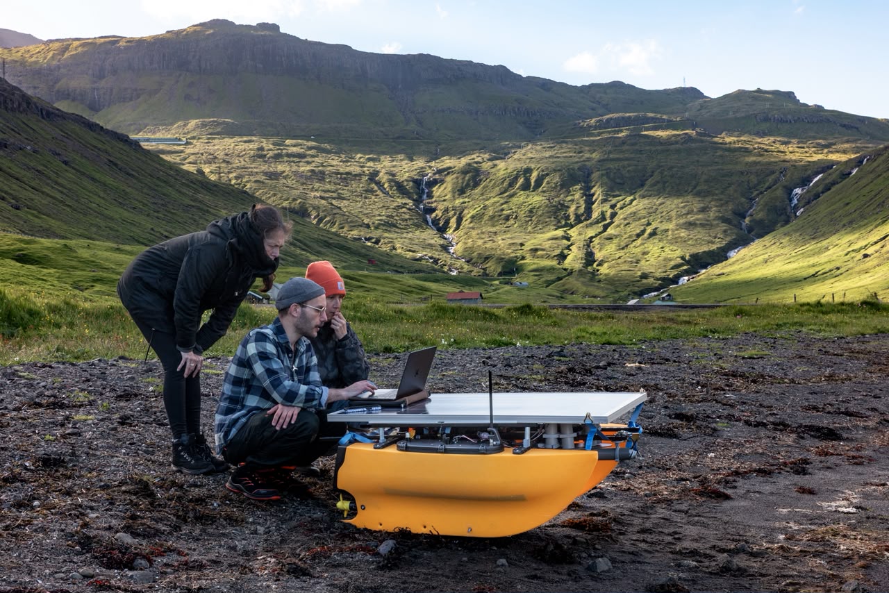 L'équipe BIUTOPIC accroupie autour d'HYDRO-01 et d'un ordinateur portable, dans une vallée des îles Féroé striée de cascades